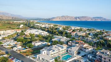 An aerial view of a coastal residential area with modern buildings, a swimming pool, and a clear view of the sea and mountains in the background.