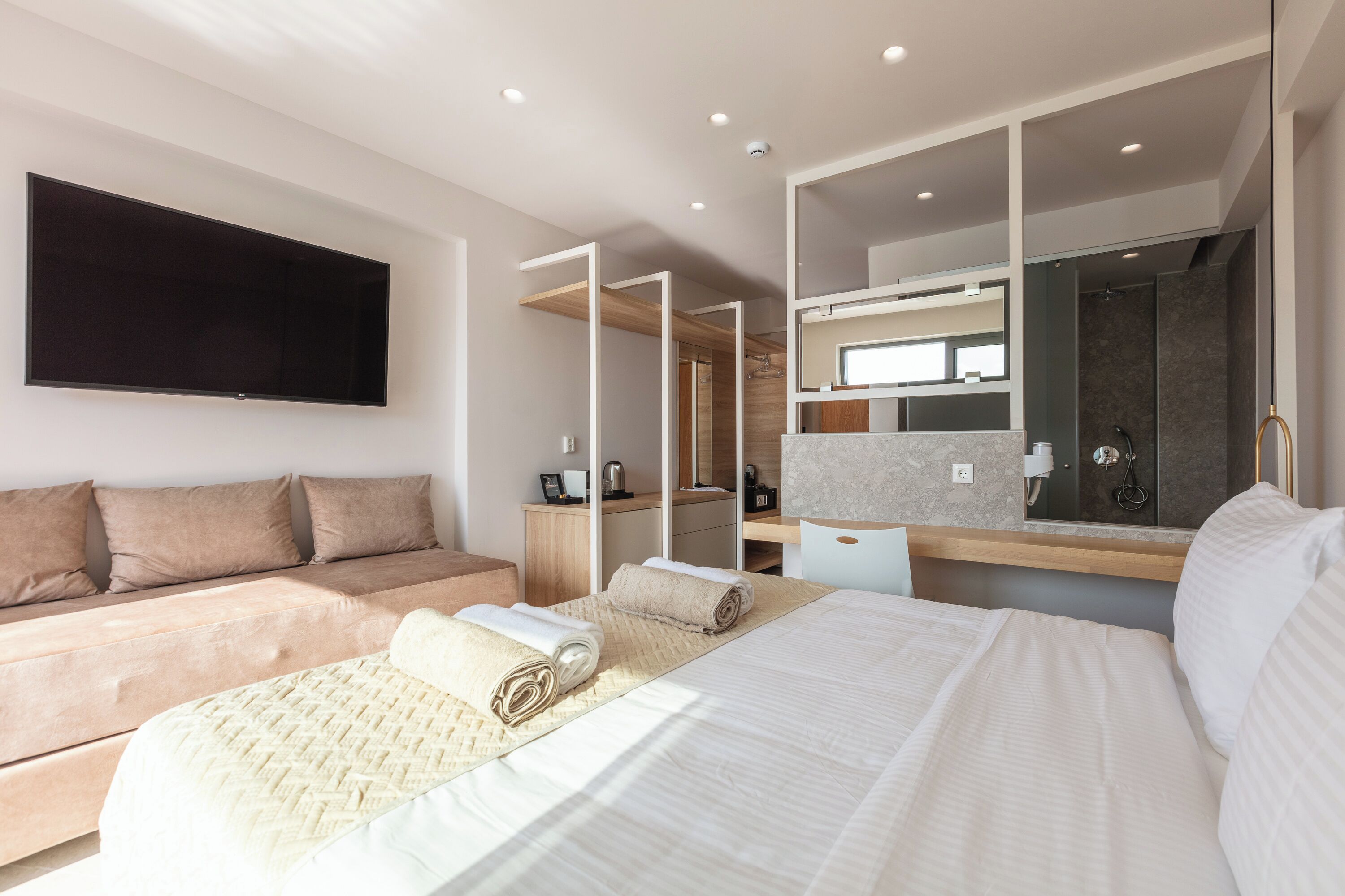 A modern hotel room with a large bed, a sofa, a flat-screen TV, a bathroom with a mirror, and a desk.
