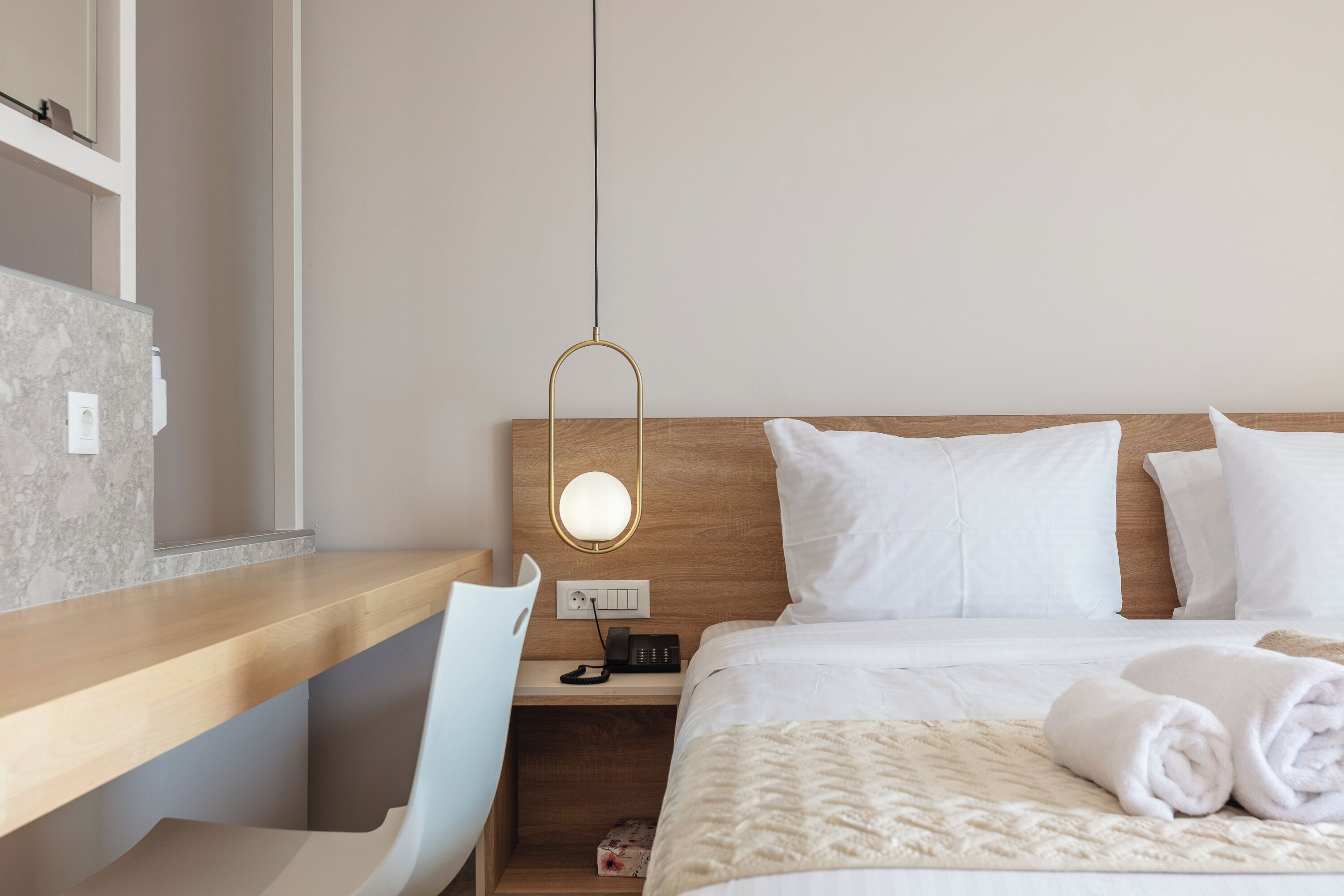 Habitación de hotel impecable con cabecera de madera, mesita de noche con lámpara, cama blanca con almohadas y una toalla enrollada sobre la cama.