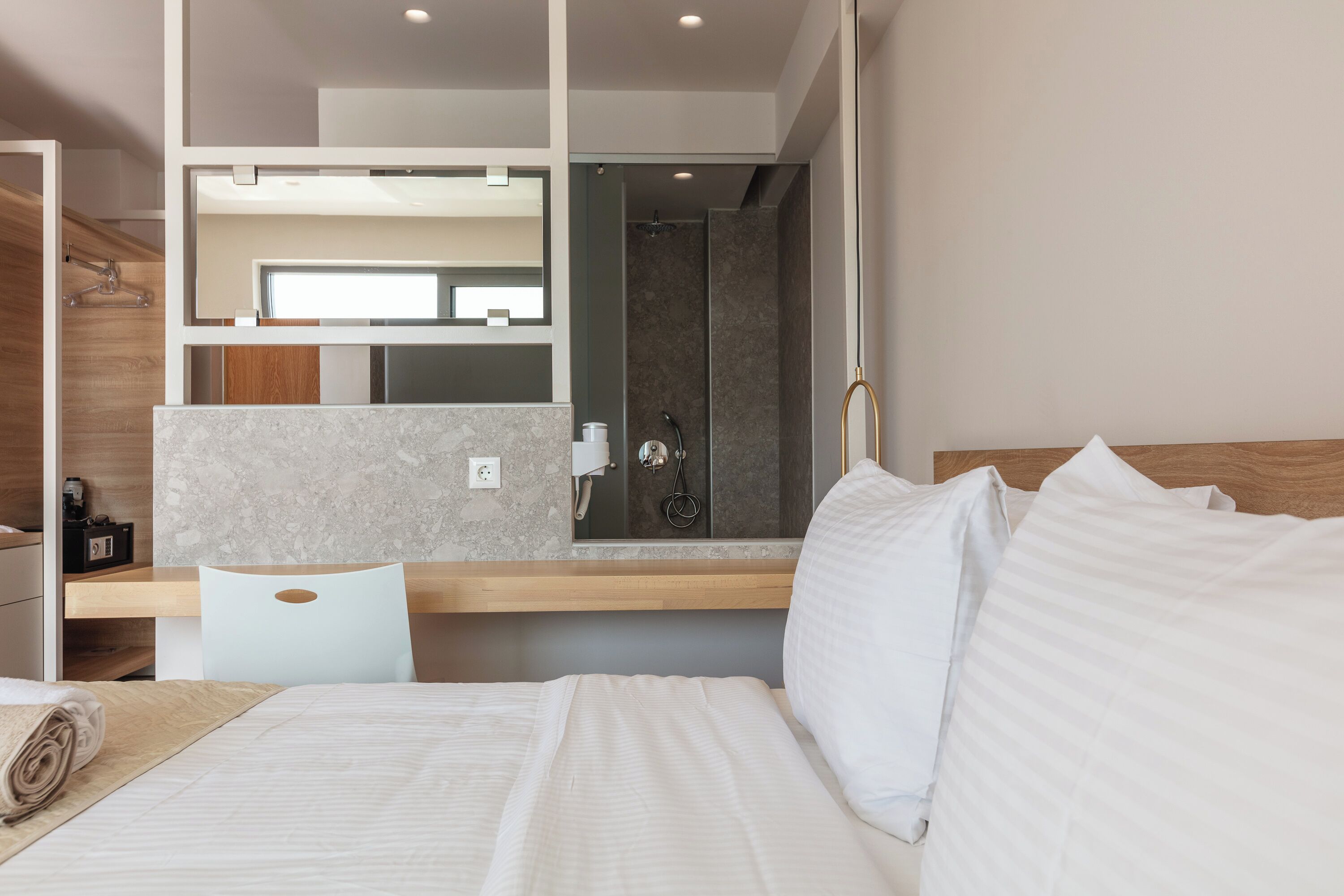 Una habitación de hotel moderna con cama, escritorio, silla, zona de ducha y un espejo grande.