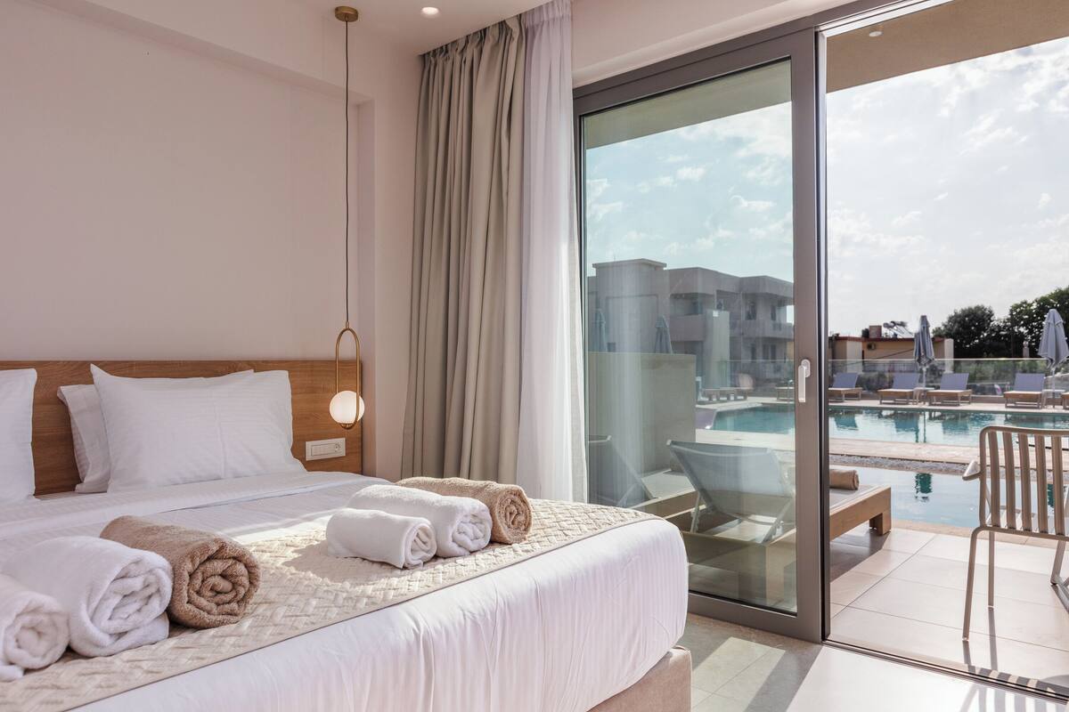 A hotel room with a large bed, a glass door leading to a pool area, and a view of the pool and buildings.