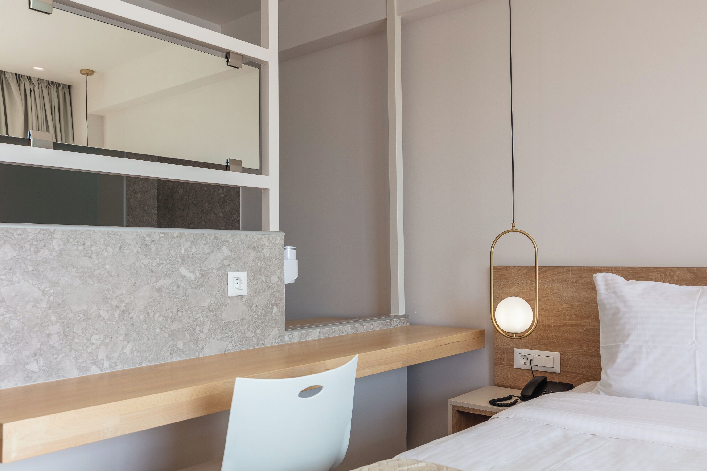A hotel room with a wooden desk, a white chair, a bed with white bedding, a nightstand with a phone, and a wall-mounted mirror.