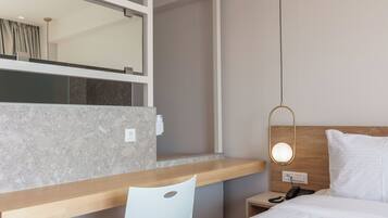 A hotel room with a wooden desk, a white chair, a bed with white bedding, a nightstand with a phone, and a wall-mounted mirror.