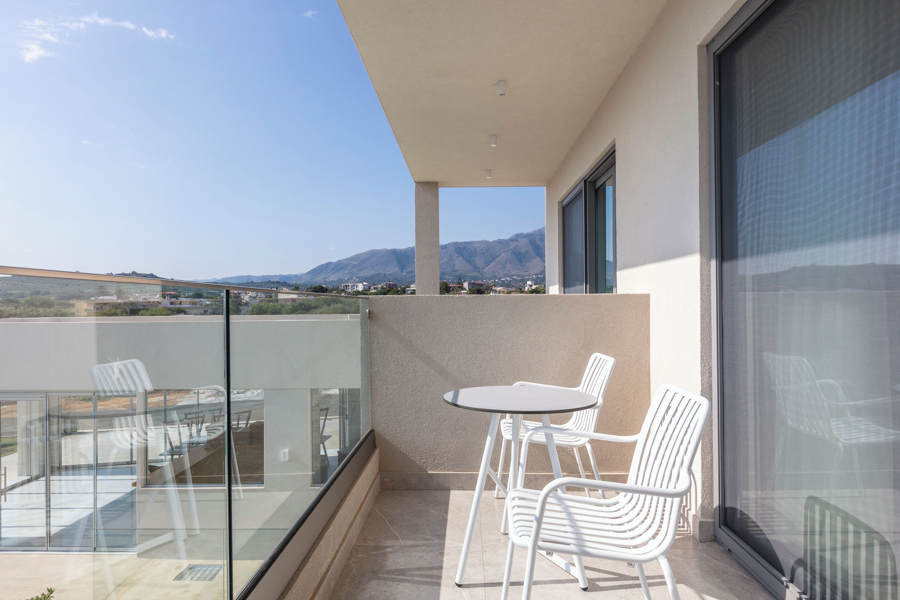 A balcony with a glass railing, white chairs, a small table, and a view of mountains and a bridge.