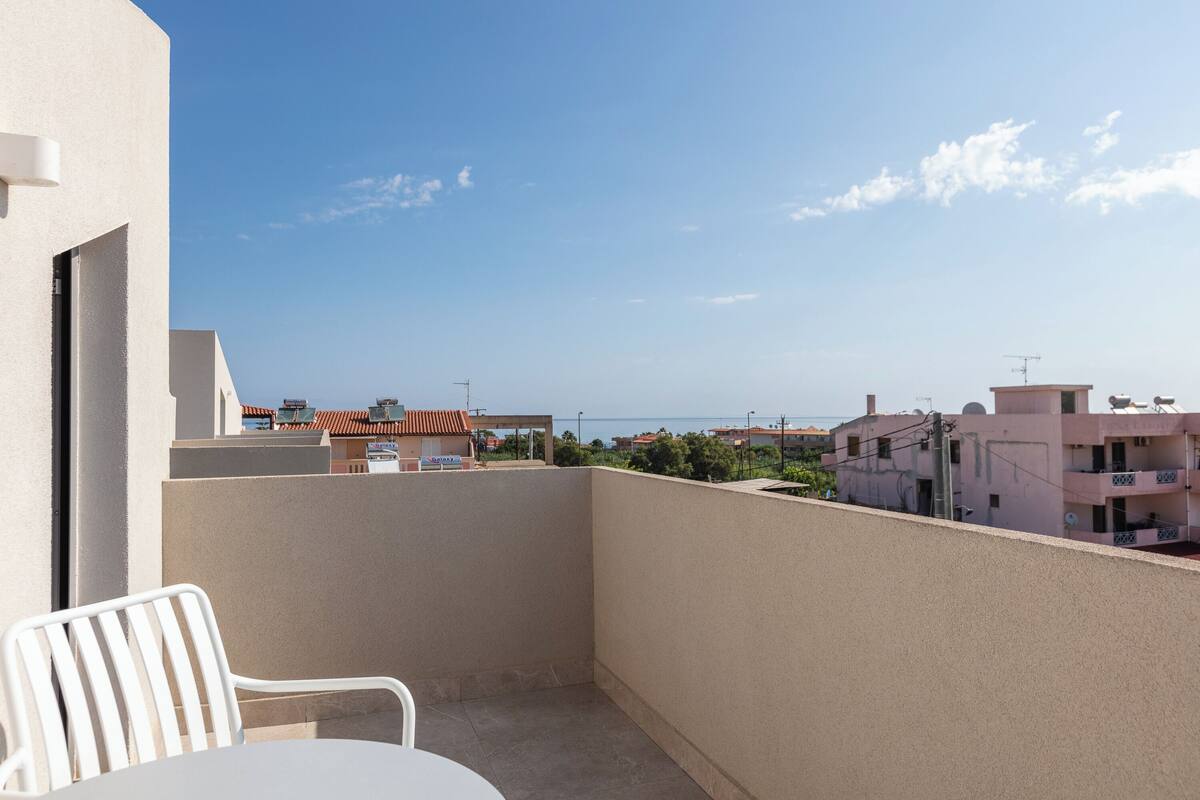 A balcony with a white chair, a table, and a view of buildings and the sea.