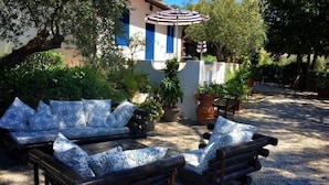 A sunlit outdoor seating area with a wooden bench and a sofa, surrounded by potted plants and a building with blue shutters. - Hotel Villa Giulia (Porto Azzurro)