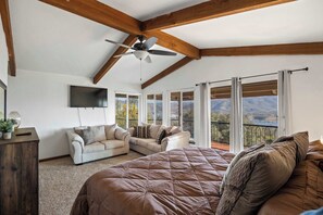 A bedroom with a large bed, a ceiling fan, a TV, and a balcony with a view of the mountains.