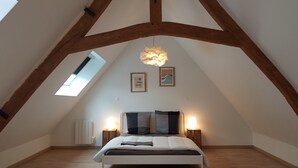 Une chambre avec un plafond en pente, des poutres en bois, un lit avec des oreillers, deux lampes de chevet et une lucarne.