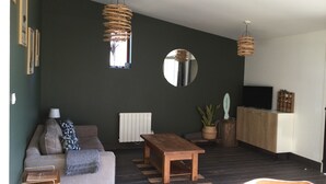 Un salon avec un canapé, une table basse et un téléviseur posé sur un meuble en bois. On y trouve deux lampes suspendues en osier, un miroir et une plante en pot.