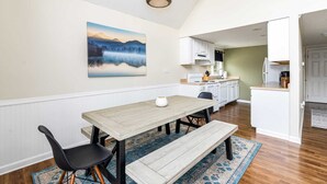 A dining area with a wooden table, black chairs, and a bench. There is a kitchen in the background with white cabinets and a stove. - Hot Tub, Fire Pit, Deck, Grill, WiFi, Dog-Friendly (Gerrardstown)