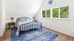 Une chambre à coucher avec un lit, un tapis bleu à motifs, une fenêtre donnant sur des arbres et un ventilateur de plafond.