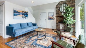 Un salon avec un canapé bleu, une table basse, un tapis à motifs, une cheminée en pierre et une vue sur l’extérieur.