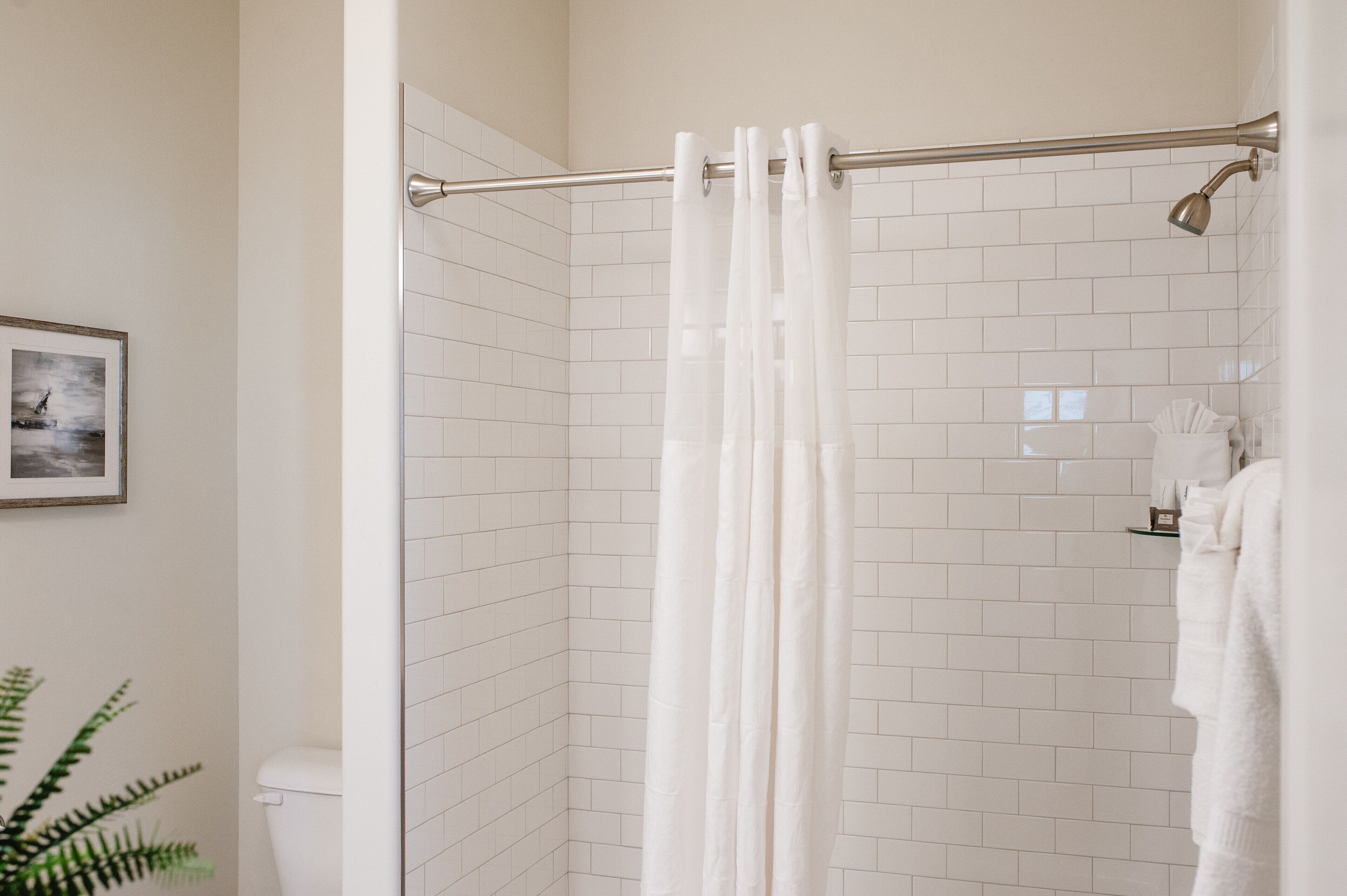 Une salle de bain avec des murs carrelés de carreaux blancs, une douche avec un rideau, des toilettes et un tableau encadré au mur.