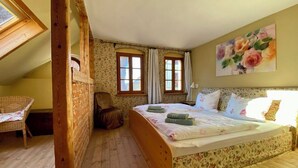 Un dormitorio con una cama de madera, una silla, un ventanal con cortinas y un cuadro floral en la pared.