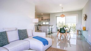 A modern living room with a white sofa, a dining area with a glass table, and a kitchen in the background. - Spacious Retreat- Pool, Gym, 3 Balconies & Parking (Playa del Carmen)