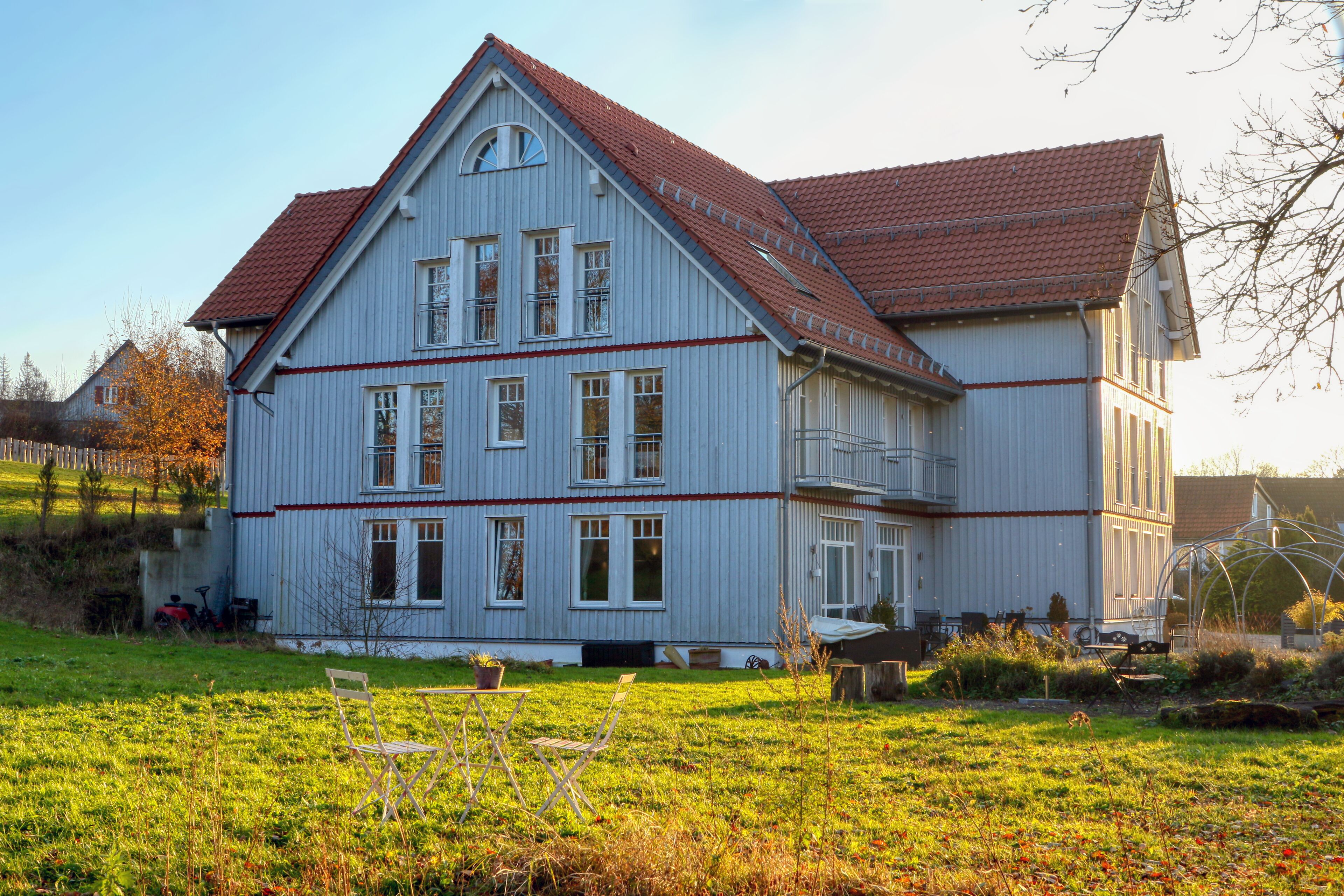 Ein großes, zweistöckiges Haus mit rotem Ziegeldach und weißem Außenputz, umgeben von einem Rasen und einem kleinen Garten.