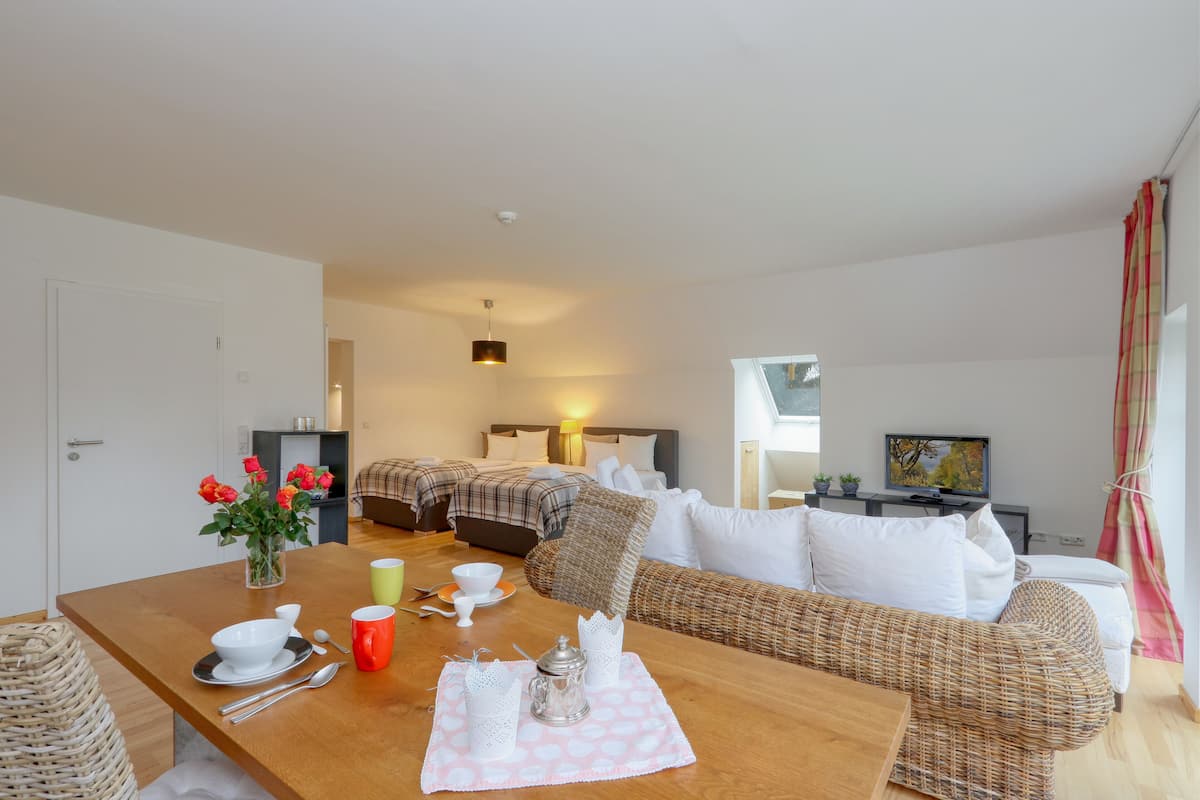 A modern hotel room with a wooden dining table set for two, a sofa, a bed with a headboard, a television, and a window with curtains.