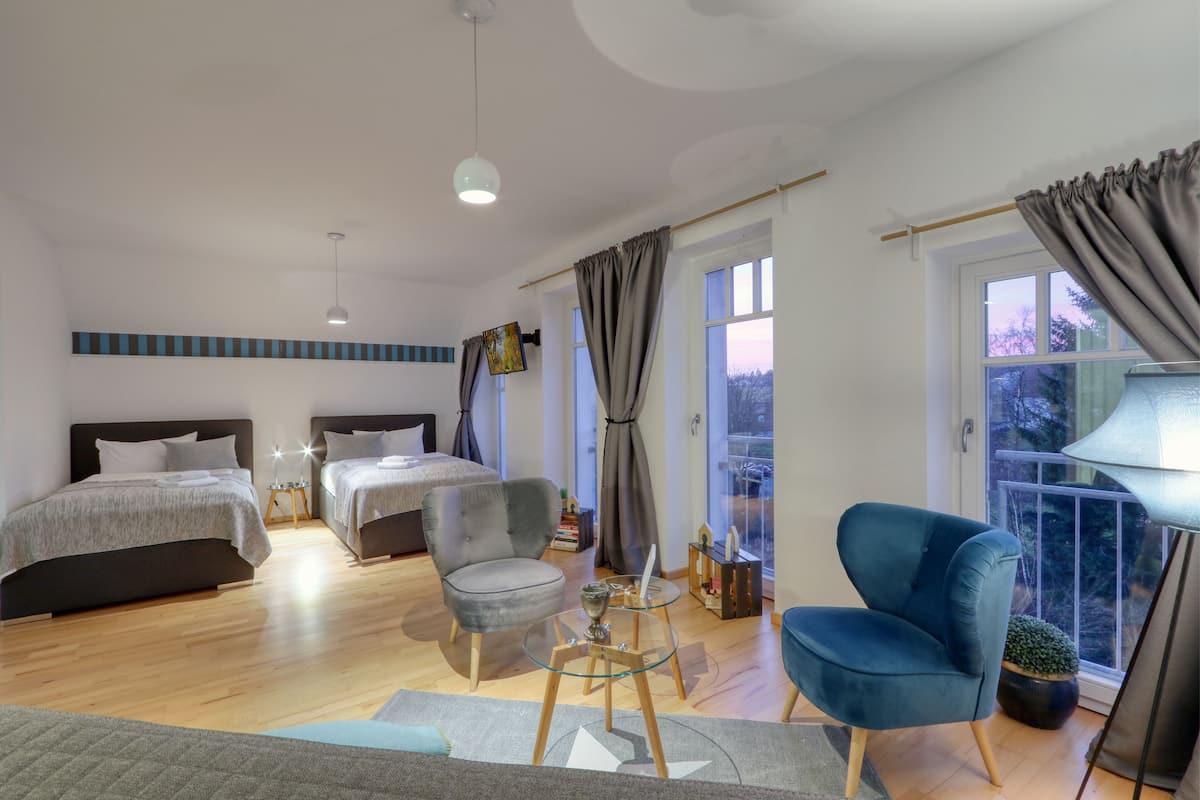 A modern bedroom with two beds, a grey armchair, a glass coffee table, and a view of the outdoors through large windows.