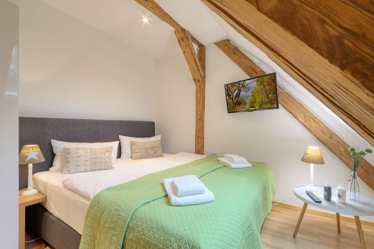 A bedroom with a bed, pillows, a green blanket, a bedside table with a lamp, a small table with a vase, and a painting on the wall.