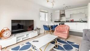 A modern living room with a flat-screen TV, a white entertainment unit, a pink armchair, and a dining area with blue chairs. - Stunning Modern Apartment in Rochester, England (Rochester)