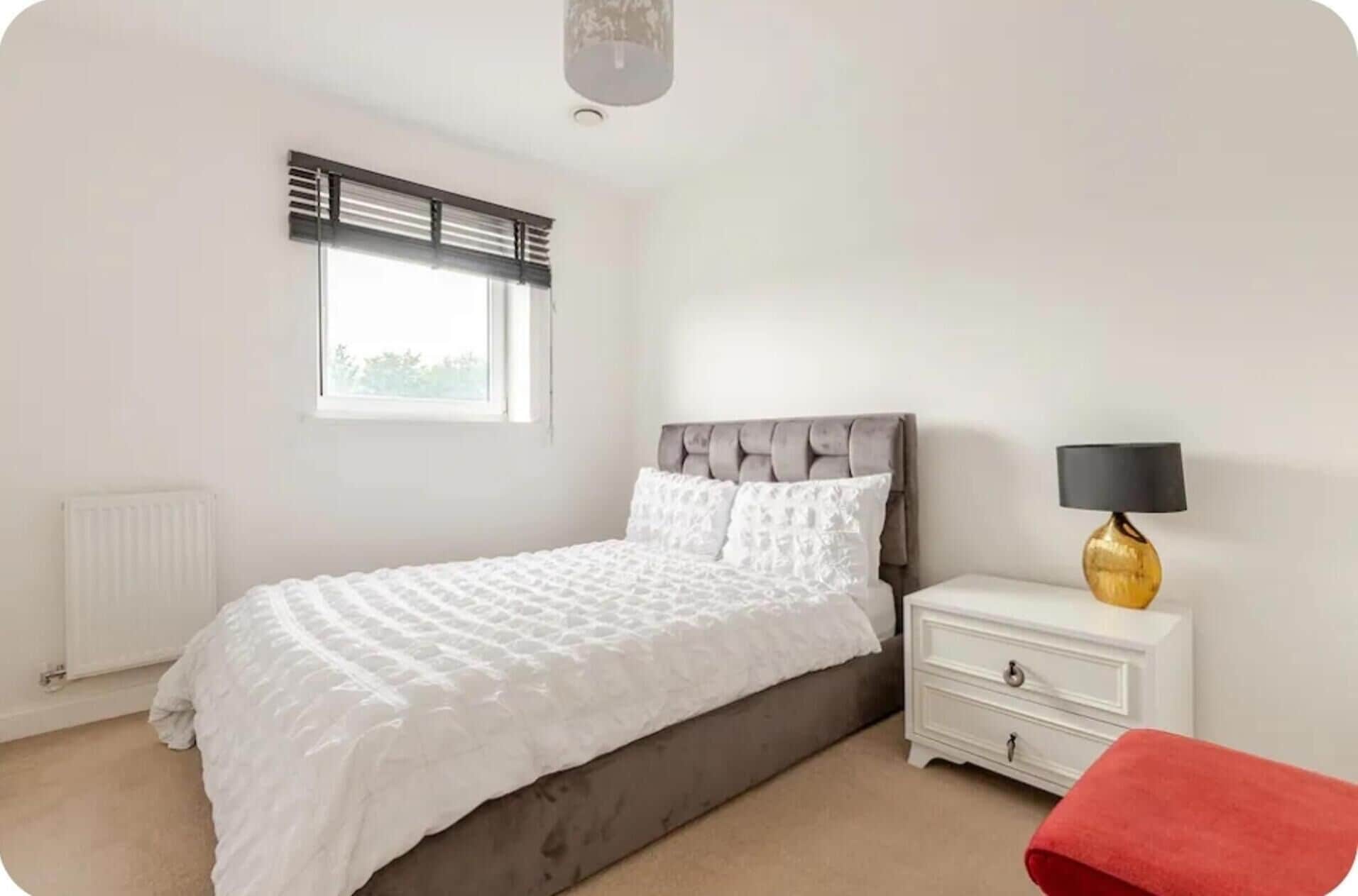 A bedroom with a bed, a bedside table with a lamp, a window with blinds, and a radiator.