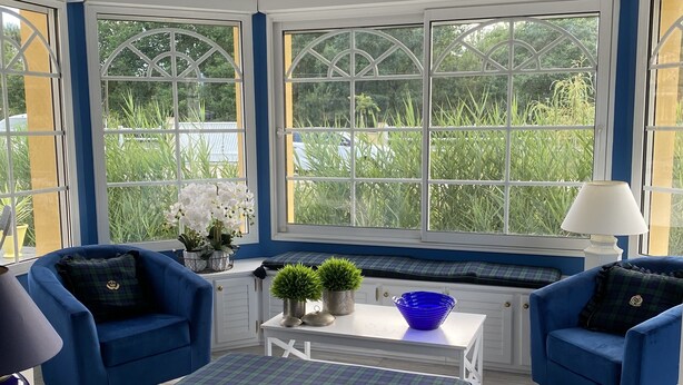 Une véranda aux murs bleus et blancs, avec deux fauteuils bleus, une table blanche et un pouf bleu.