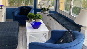 Une véranda aux murs bleus et aux boiseries blanches, comprenant un canapé bleu, une table blanche avec un saladier bleu et un banc avec une lampe.