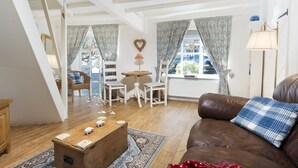 A cozy living room with a brown leather sofa, a wooden coffee table, and a patterned rug. There are two white chairs and a small wooden table with a lamp. The room has white walls, wooden beams on the ceiling, and large windows with patterned curtains. - The Old Dairy - One Bedroom House, Sleeps 2 (Clappersgate)