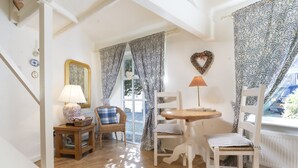 A cozy room with a wooden floor, a round table, two wicker chairs, a blue and white patterned curtain, a heart-shaped decoration, and a lamp on the table. - The Old Dairy - One Bedroom House, Sleeps 2 (Clappersgate)