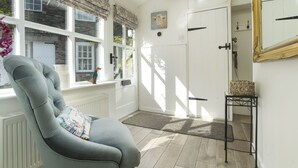 A room with a grey upholstered chair, a white door, a window with a floral valance, and a small table with a basket. - The Old Dairy - One Bedroom House, Sleeps 2 (Clappersgate)