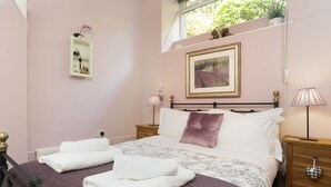 A bedroom with a bed, two bedside tables, a nightstand, a window with a view of trees, and a shelf with decorative items. - The Old Dairy - One Bedroom House, Sleeps 2 (Clappersgate)