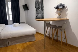 Une chambre d’hôtel avec un lit, une petite table en bois et deux chaises, une plante en pot et un téléviseur fixé au mur.