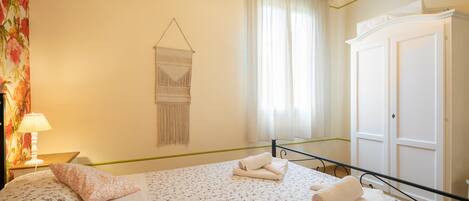 Un dormitorio con una cama, una mesita de noche con lámpara, un tapiz de macramé en la pared y un armario.