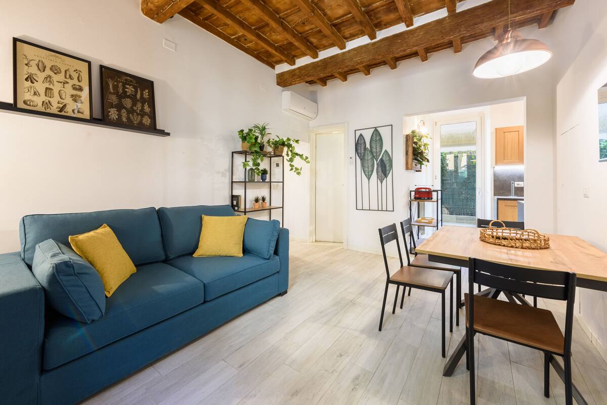 A modern living room with a blue sofa, wooden dining table, and chairs. There are framed botanical prints on the wall and a plant on a shelf.