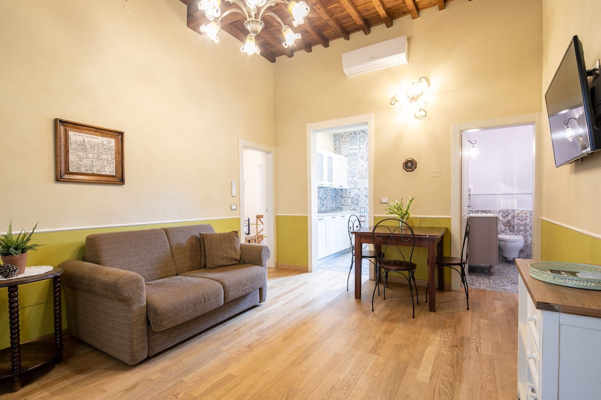 A cozy living room with a sofa, a dining table with chairs, a TV mounted on the wall, and a kitchen area visible through an open door.