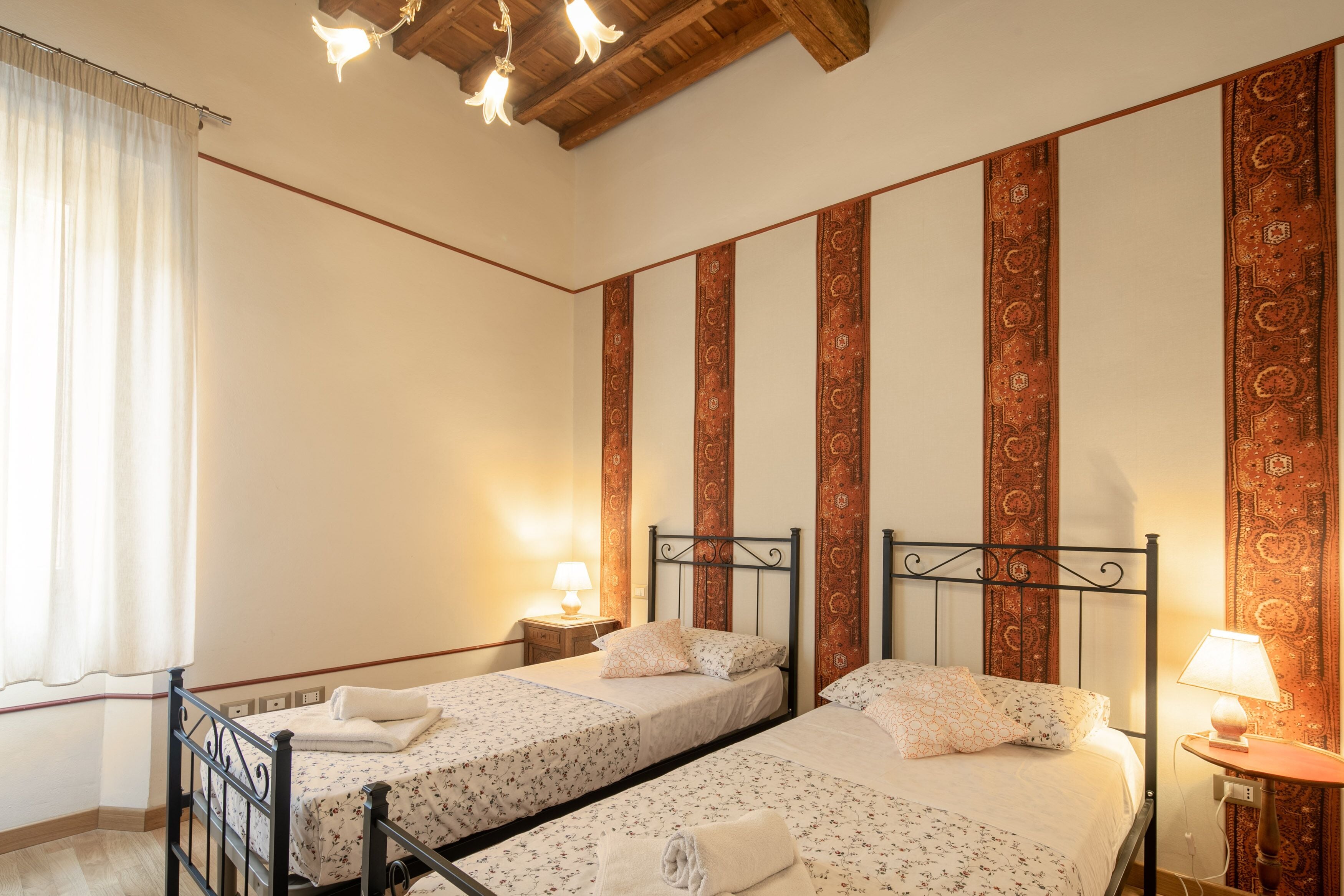 Habitación de hotel con dos camas individuales, techo de madera, cortinas estampadas y una mesita de noche con lámpara.