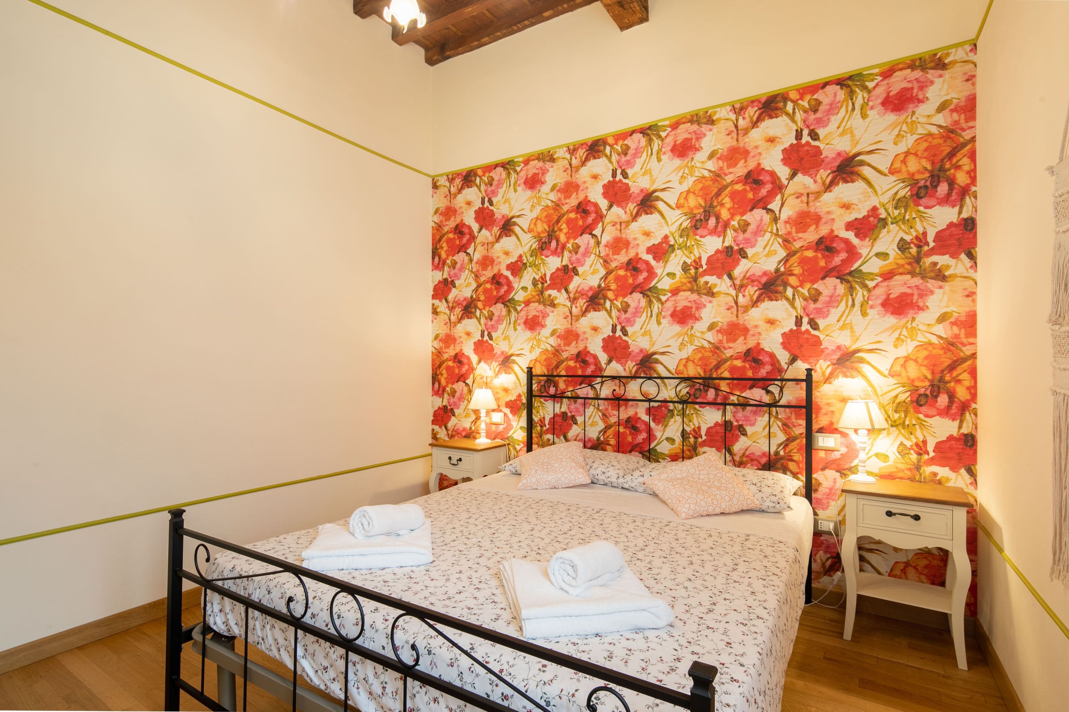 A bedroom with a floral wallpaper, a metal bed frame, two bedside tables with lamps, and neatly folded towels on the bed.