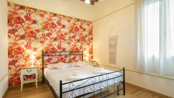 Une chambre à coucher avec un papier peint à motifs floraux, un lit avec un cadre en métal, une table de chevet, une lampe et une fenêtre avec des rideaux.