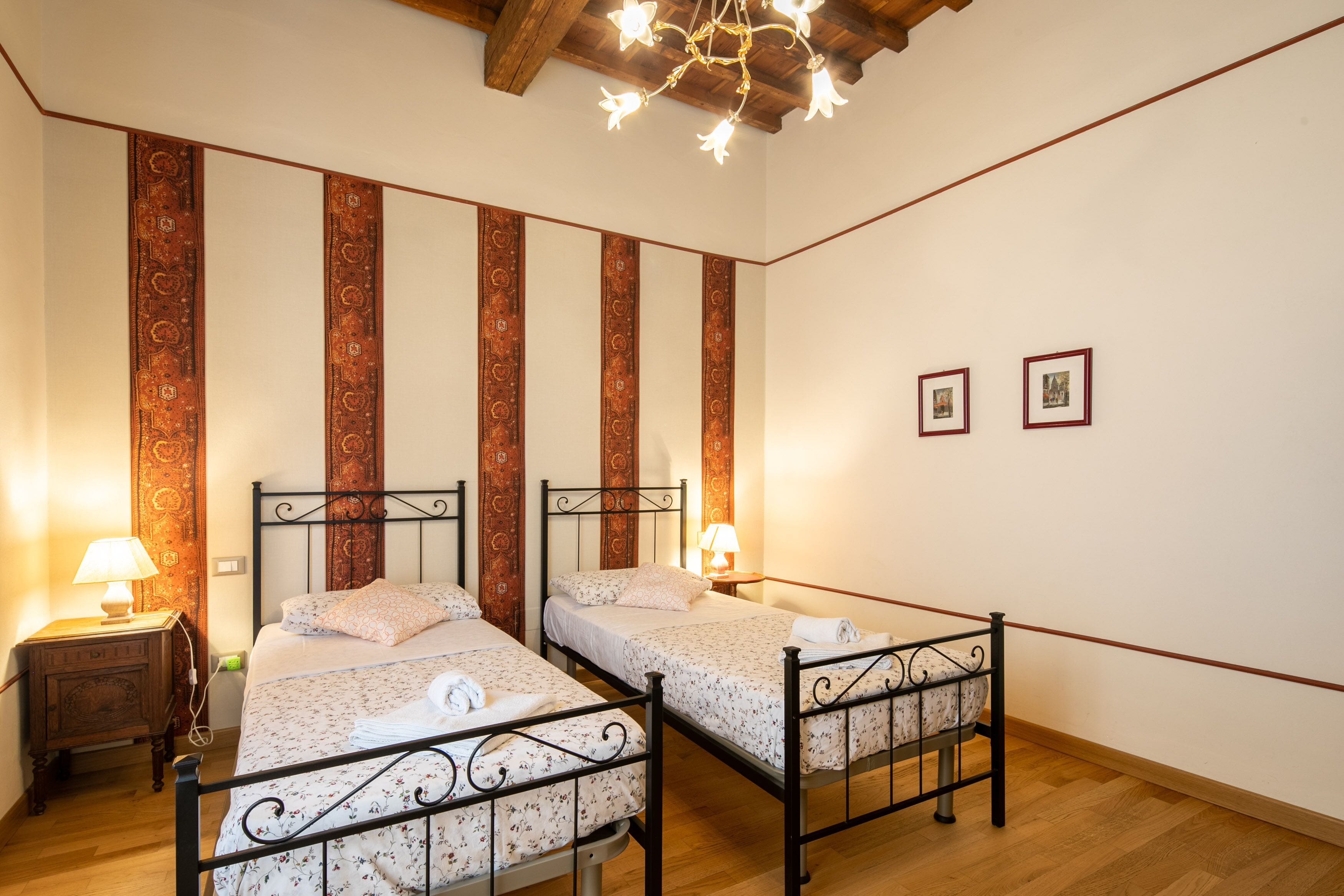 A room with two beds, wooden floors, patterned curtains, and a chandelier.
