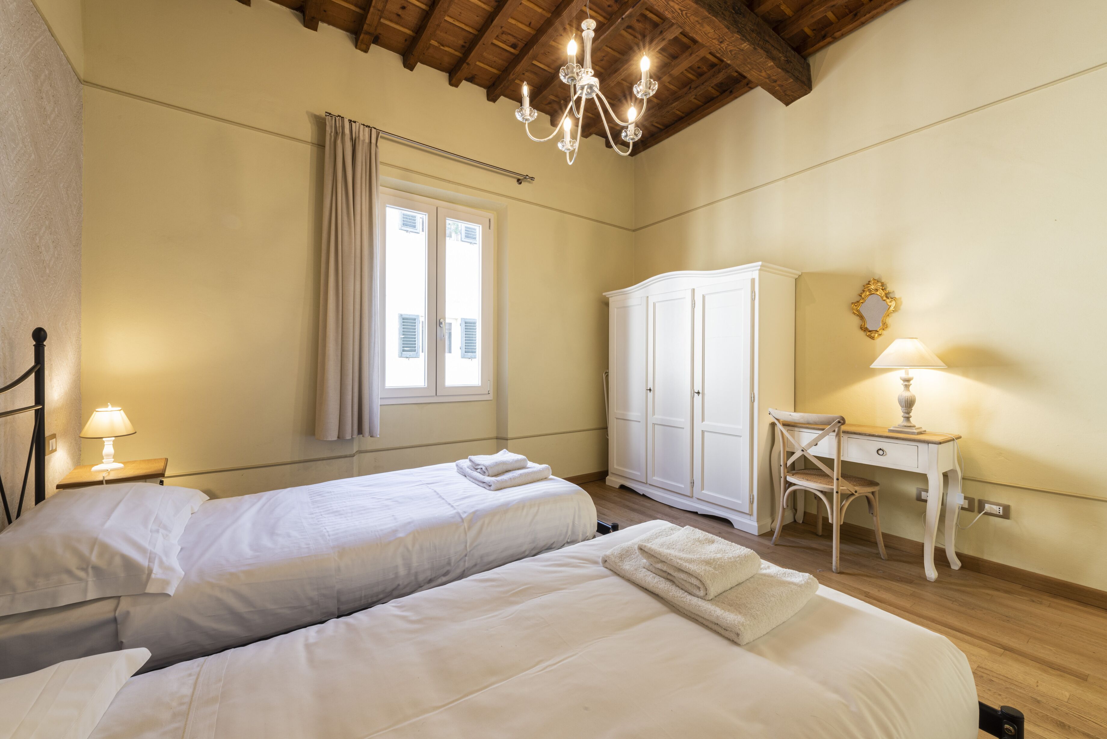 A bedroom with two beds, a desk, a chair, a wardrobe, and a chandelier.