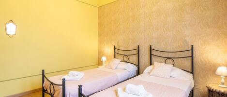 A room with two beds, wooden floors, patterned wallpaper, and a small bedside table with a lamp.