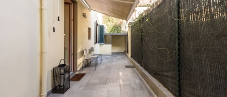 A narrow outdoor corridor with a tiled floor, a covered seating area, and a metal fence.