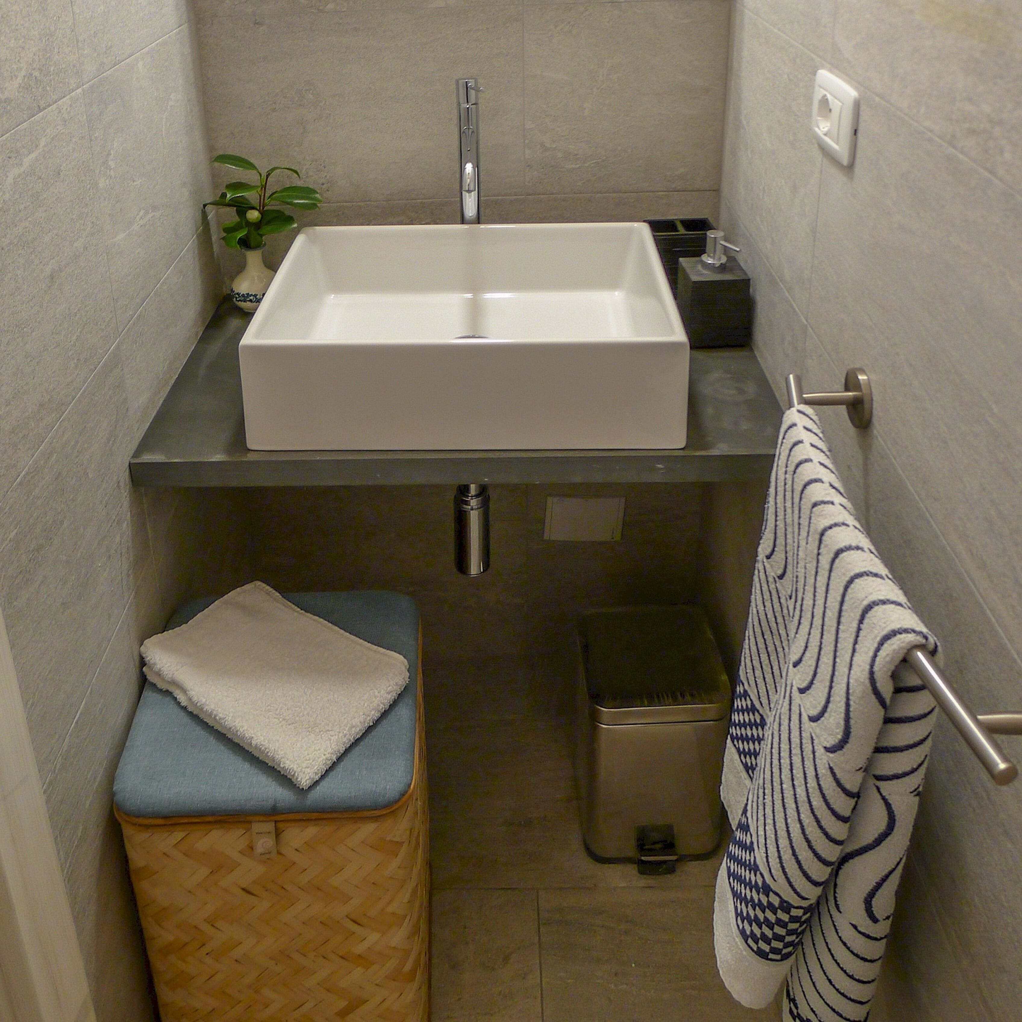 A bathroom with a rectangular sink, a small trash bin, a towel rack, and a plant.