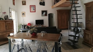 Amplia sala de estar con mesa de comedor de madera, sillas traslúcidas, sofá y escalera.