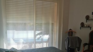 Un dormitorio con cama, dos almohadas, una silla, una mesa y una ventana con persianas.
