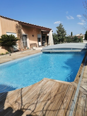 Une piscine rectangulaire avec une terrasse en bois, une maison beige avec un toit rouge et un ciel bleu clair.