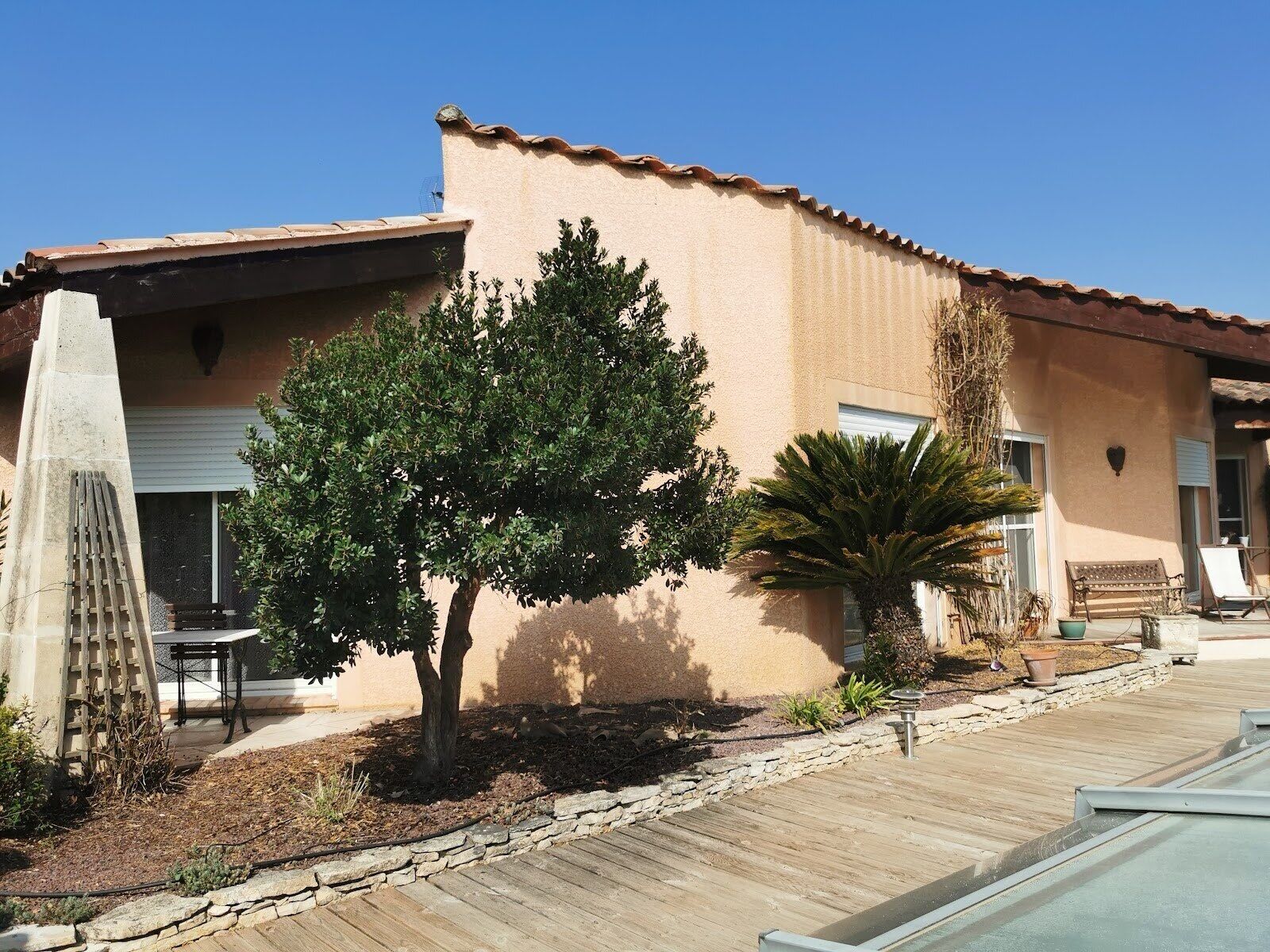 A house with a tiled roof, a patio with a wooden deck, and a small garden with a tree and a palm plant.