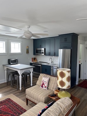 Une cuisine moderne avec des meubles de couleur bleu foncé, un réfrigérateur en acier inoxydable et un four à micro-ondes. On y trouve un coin repas avec une table blanche et deux tabourets de bar. Un coussin décoratif à motifs est posé sur un canapé beige, et une lampe se trouve sur une table d’appoint.