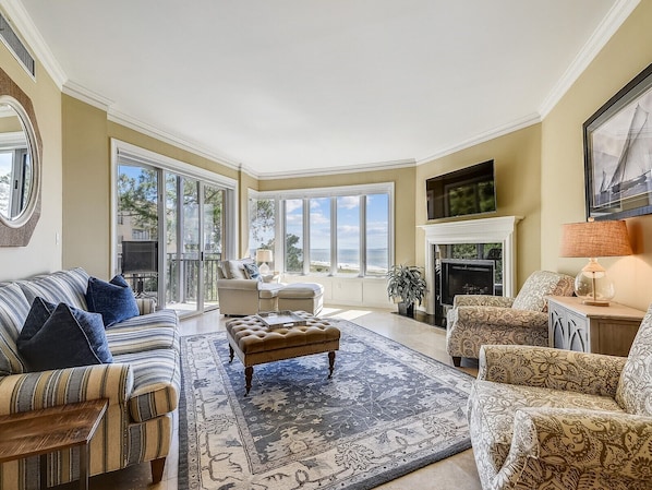 A spacious living room with a large window overlooking the beach, a fireplace, and a cozy seating area.