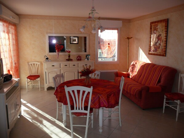 A dining area with a round table, white chairs, and a red tablecloth. There is a sofa with a striped cushion and a painting on the wall.